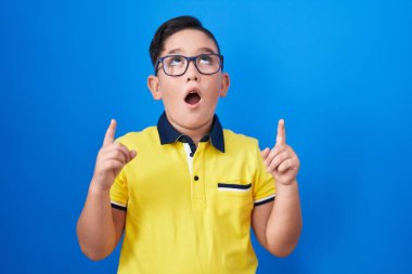 Young hispanic kid standing over blue background amazed and surprised looking up and pointing with fingers and raised arms. 