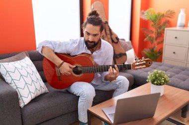 Young hispanic man having online guitar class sitting on sofa at home