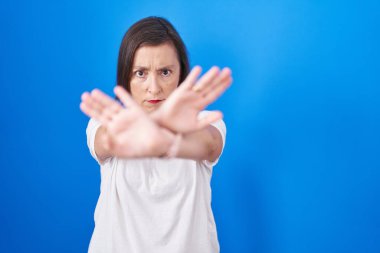 Orta yaşlı İspanyol bir kadın mavi arka planda duruyor. Reddedilme ifadesi. Kollarını kavuşturup, avuç içlerine bakıyor. 