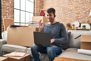 Yeni evinde dizüstü bilgisayarla çalışan genç İspanyol bir adam parmaklarıyla imza atıyor, gülümseyerek mükemmel bir sembol oluşturuyor. 