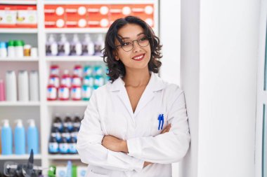 Young beautiful hispanic woman pharmacist smiling confident standing with arms crossed gesture at pharmacy