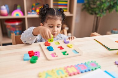 Adorable hispanic girl playing with maths puzzle game sitting on table at kindergarten