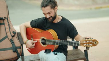Young hispanic man musician playing classical guitar sitting on bench at street