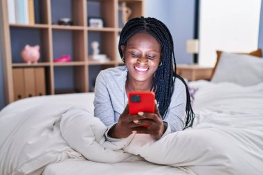 African american woman using smartphone lying on bed at bedroom