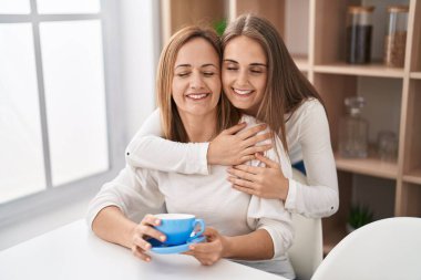 Two women mother and daughter hugging each other drinking coffee at home