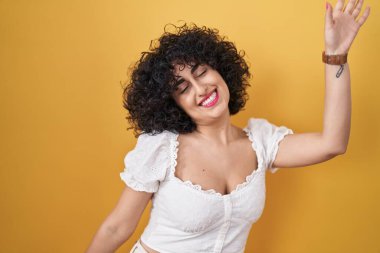 Sarı arka planda kıvırcık saçlı genç esmer kadın dans ediyor mutlu ve neşeli, hareketli ve kendinden emin müzik dinliyor. 
