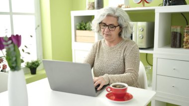 Middle age woman with grey hair using laptop sitting on table at home