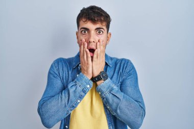 Young hispanic man standing over blue background afraid and shocked, surprise and amazed expression with hands on face 