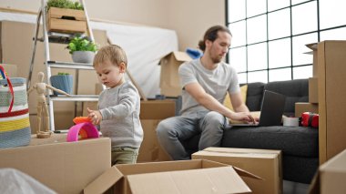 Baba ve oğul yeni evlerinde dizüstü bilgisayar kullanarak karton kutuları açıyor.