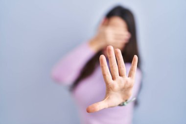 Genç esmer kadın mavi arka planda duruyor ellerle gözleri örtüyor ve üzgün ve korku ifadeleriyle dur hareketi yapıyor. utangaç ve olumsuz kavram. 