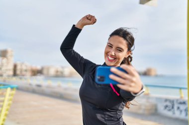 Genç İspanyol kadın güçlü bir jest yapıyor. Deniz kenarındaki akıllı telefonun yanında selfie çekiyor.