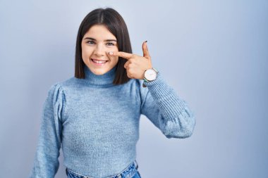 Mavi arka planda dikilen genç İspanyol kadın, parmakları burnuna ve yüzüne dönük, neşeli bir şekilde gülümsüyor. güzellik konsepti 