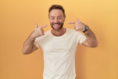 Sarı arka planda sakallı, neşeli gülümseyen, parmakları ve ağzıyla işaret eden orta yaşlı bir adam. diş sağlığı kavramı. 