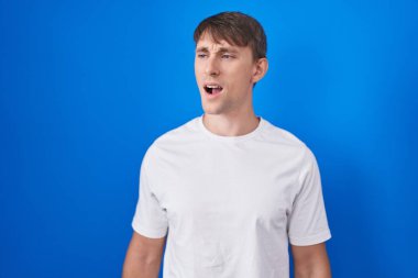 Kafkasyalı sarışın adam mavi arka planda kızgın ve kızgın bağırıyor sinirli ve öfkeli, öfkeyle bağırıyor. öfke ve agresif konsept. 