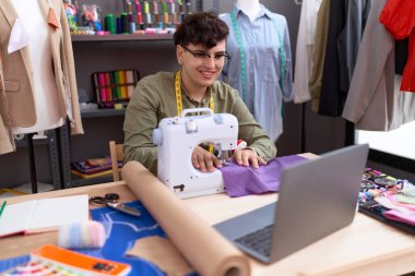 Non binary man tailor using sewing machine and laptop at atelier