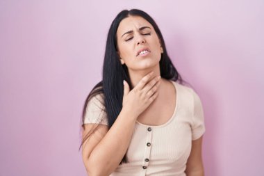 Pembe arka planda duran genç İspanyol kadın ağrılı boynuna dokunuyor, boğazı ağrıyor grip, mantar ve enfeksiyon kapmış. 