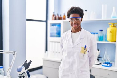 Laboratuvarda dikilen bilim adamı üniforması giyen Afrikalı Amerikalı kadın.