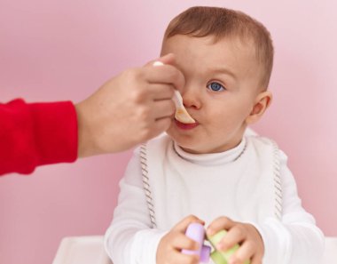 Sevimli sarışın çocuk, sandalyeye oturmuş pembe arka planda yemek yiyor.
