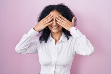 Hispanic woman with curly hair standing over pink background covering eyes with hands smiling cheerful and funny. blind concept. 