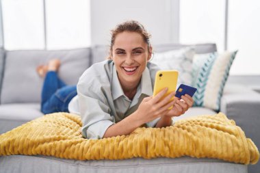 Akıllı telefon ve kredi kartı kullanan genç bir kadın evdeki kanepede yatıyor.
