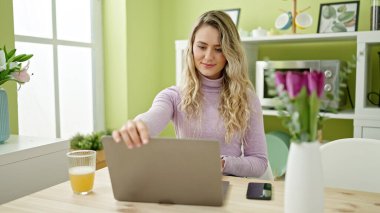 Genç sarışın kadın gülerek yemek odasında dizüstü bilgisayar açıyor.