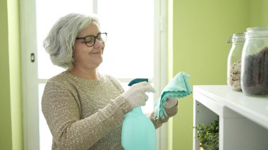 Middle age woman with grey hair holding cloth and sprayer at home