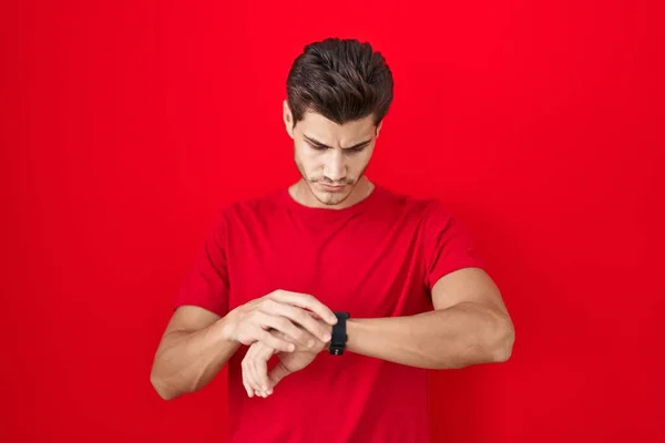 Młody Hiszpan Stojący Nad Czerwonym Tłem Sprawdzający Czas Zegarku Zrelaksowany — Zdjęcie stockowe