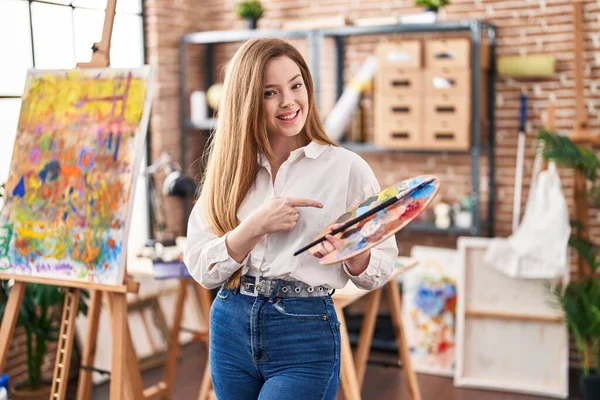 艺术工作室里年轻的高加索女人牵着调色板 高兴地微笑着 手指手画脚 — 图库照片