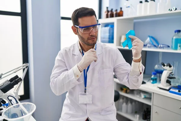 stock image Young hispanic man with beard working at scientist laboratory holding blue ribbon serious face thinking about question with hand on chin, thoughtful about confusing idea 