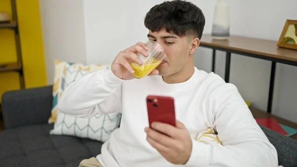 Молодой Латиноамериканец Использующий Смартфон Пьет Апельсиновый Сок Дома — стоковое фото