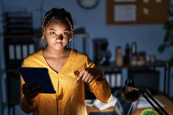 身穿辫子的非洲裔美国妇女深夜在办公室里工作 平板电脑指向下看去既悲伤又沮丧 用手指指示方向 既不快乐又沮丧 — 图库照片