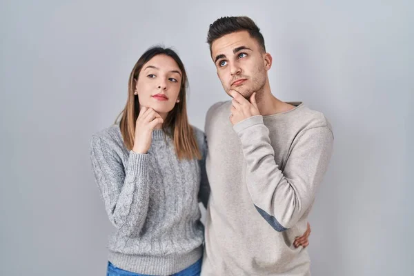 若いヒスパニック系のカップルの白の背景に手をあごの質問については 集中的な表現を考えて立っている 思慮深い顔で笑顔 疑わしい概念 — ストック写真
