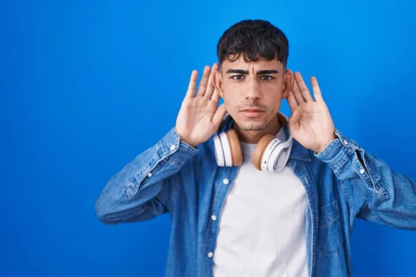 Homem Hispânico Jovem Sobre Fundo Azul Tentando Ouvir Ambas Mãos — Fotografia de Stock