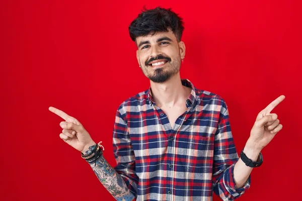 髭を生やした若いヒスパニック系の男性が 自信を持ってさまざまな方向に指を指して笑顔で赤い背景に立っている 広告のコピースペース — ストック写真