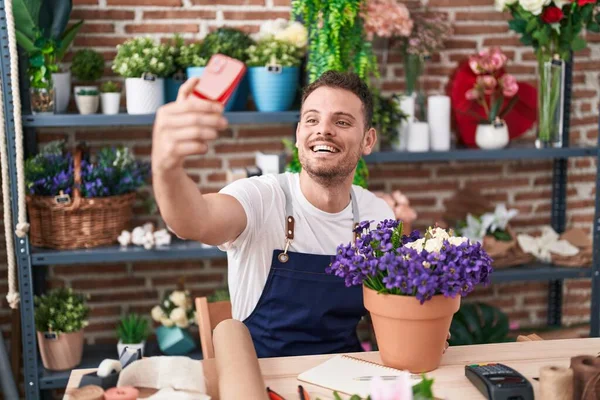 Young hispanic man florist make selfie by smartphone sitting on table at florist