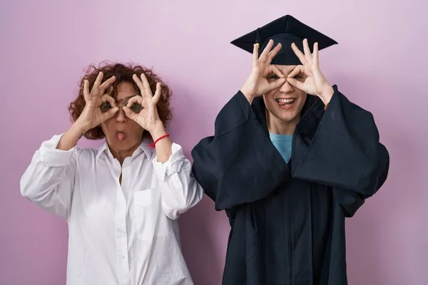 卒業キャップと式用のローブを着たヒスパニック系の母親と娘は 双眼鏡が舌を突き出しているようなOkジェスチャーをして 目は指を見ています クレイジーな表現 — ストック写真