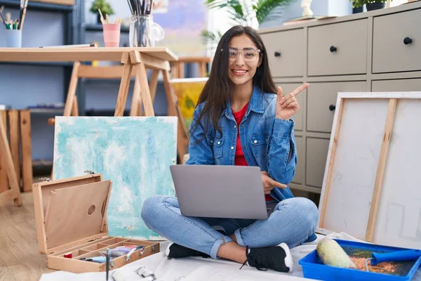 年轻的少女坐在艺术工作室里 用笔记本电脑微笑着 手指手画脚地指向旁边 — 图库照片