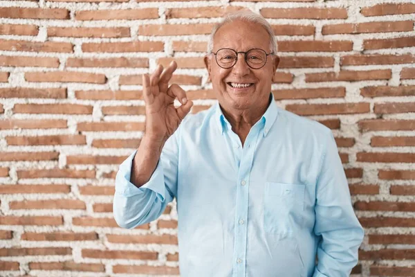 stock image Senior man with grey hair standing over bricks wall smiling positive doing ok sign with hand and fingers. successful expression. 