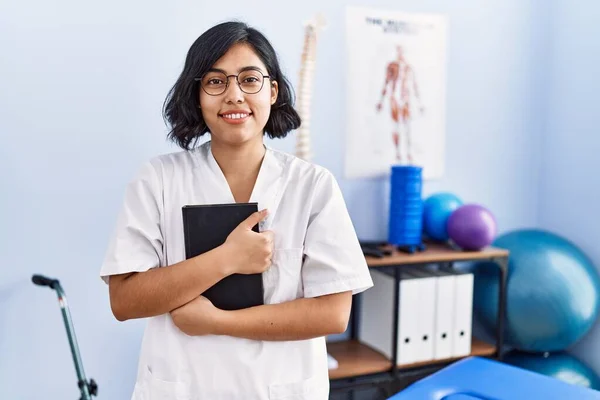 Young Latin Woman Wearing Physiotherapist Uniform Hugging Book Physiotherapy Clinic — Stock Photo, Image