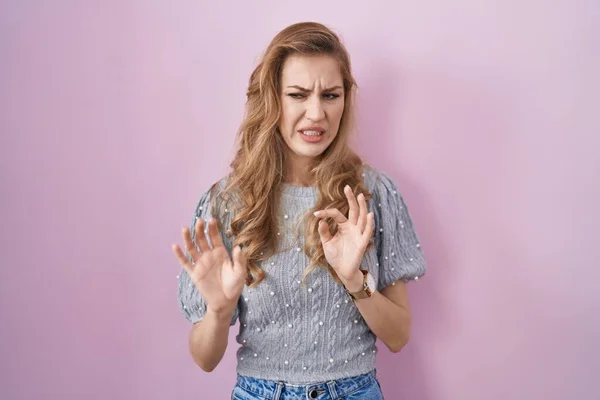 Beautiful blonde woman standing over pink background disgusted expression, displeased and fearful doing disgust face because aversion reaction.