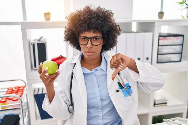 Black doctor woman with curly hair holding green apple with angry face, negative sign showing dislike with thumbs down, rejection concept