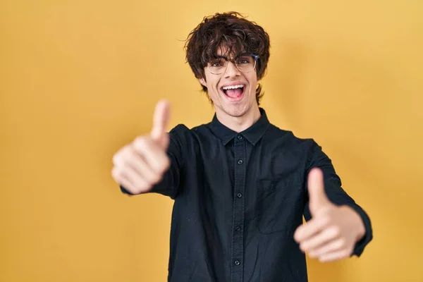年轻男子戴着黄色背景的眼镜 赞成用手做一个积极的手势 微笑着竖起大拇指 为成功感到高兴 优胜手势 — 图库照片