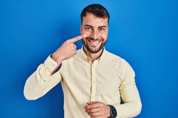 ハンサムなヒスパニック系の男性の青い背景の顔と鼻に手の指を指して立って 陽気な笑顔 美の概念 — ストック写真