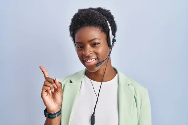 African american woman wearing call center agent headset with a big smile on face, pointing with hand finger to the side looking at the camera.
