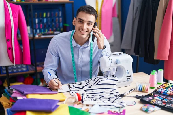 Young Hispanic Man Tailor Talking Smartphone Writing Notebook Sewing Studio — Stockfoto