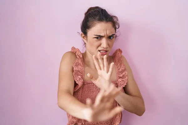 Young teenager girl standing over pink background disgusted expression, displeased and fearful doing disgust face because aversion reaction. with hands raised