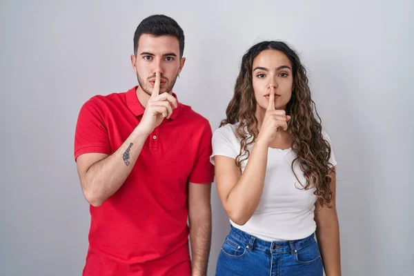 若いヒスパニック系のカップル孤立した背景の上に唇の上に指で静かにするように求めて立っている 沈黙と秘密の概念 — ストック写真