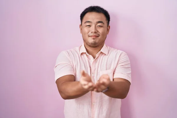 ピンクの背景の上に立つ中国の若者が手の手のひらで一緒に受信またはジェスチャーを与える笑顔 保持し保護する — ストック写真
