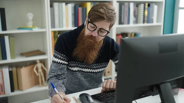 Young redhead man student using computer writing on notebook at library university