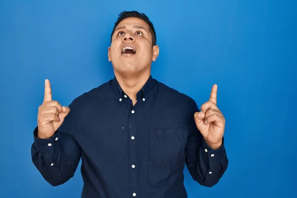 Латиноамериканец Стоящий Синем Фоне Поражен Удивлен Глядя Вверх Показывая Пальцами — стоковое фото